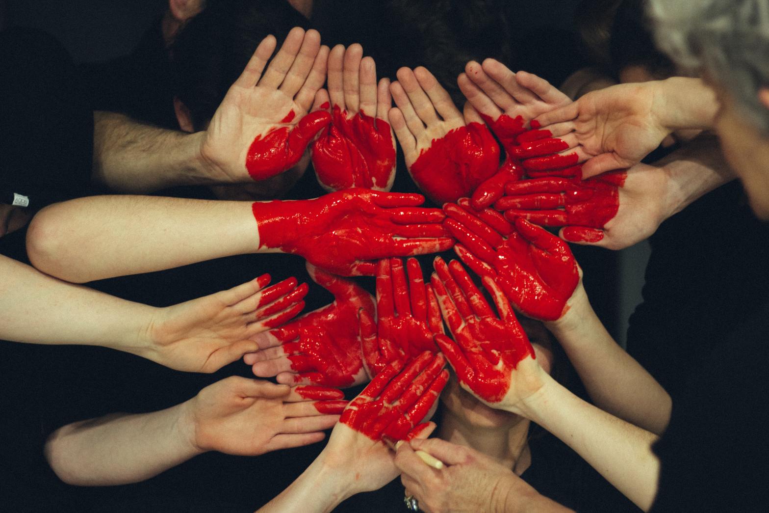 A group of people place their hands together, all palms facing outward, as another hand with a paintbrush paints a red heart shape over their hovering hands.