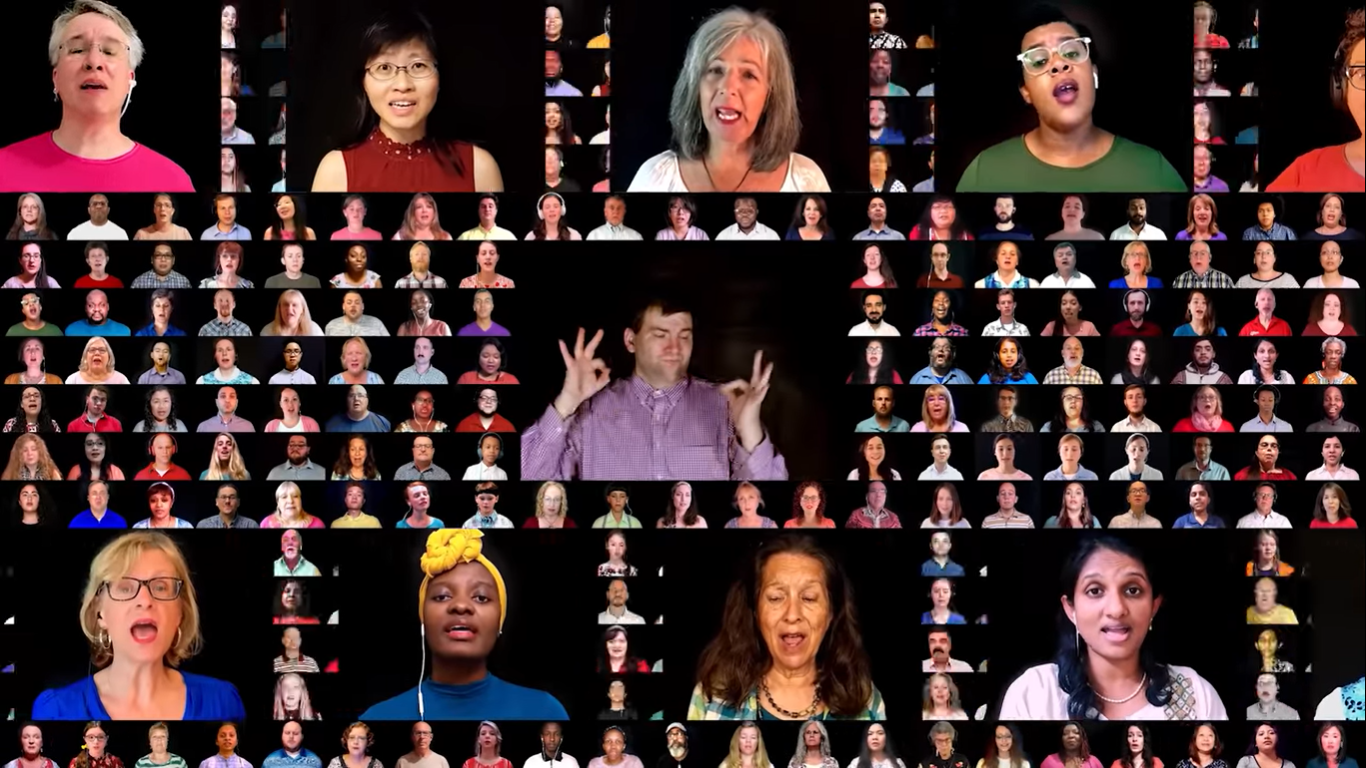 A screencap from the video "The Solid Rock." Centered is an ASL interpreter in a light purple shirt. Above and below are five horizontally scrolling singers, each wearing a shirt color corresponding to their part (see video alt text). In the background is a mosaic of singers and interpreters.